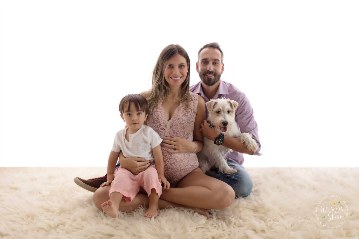 Foto feita em estúdio fotográfico localizado em Pinheiros, São Paulo, SP, para o ensaio gestante da Camila, na foto está a família toda sentada no tapete bege, a mamãe o papai o irmão mais velho e o pet da família, todos estão abraçadinhos e sorridentes.