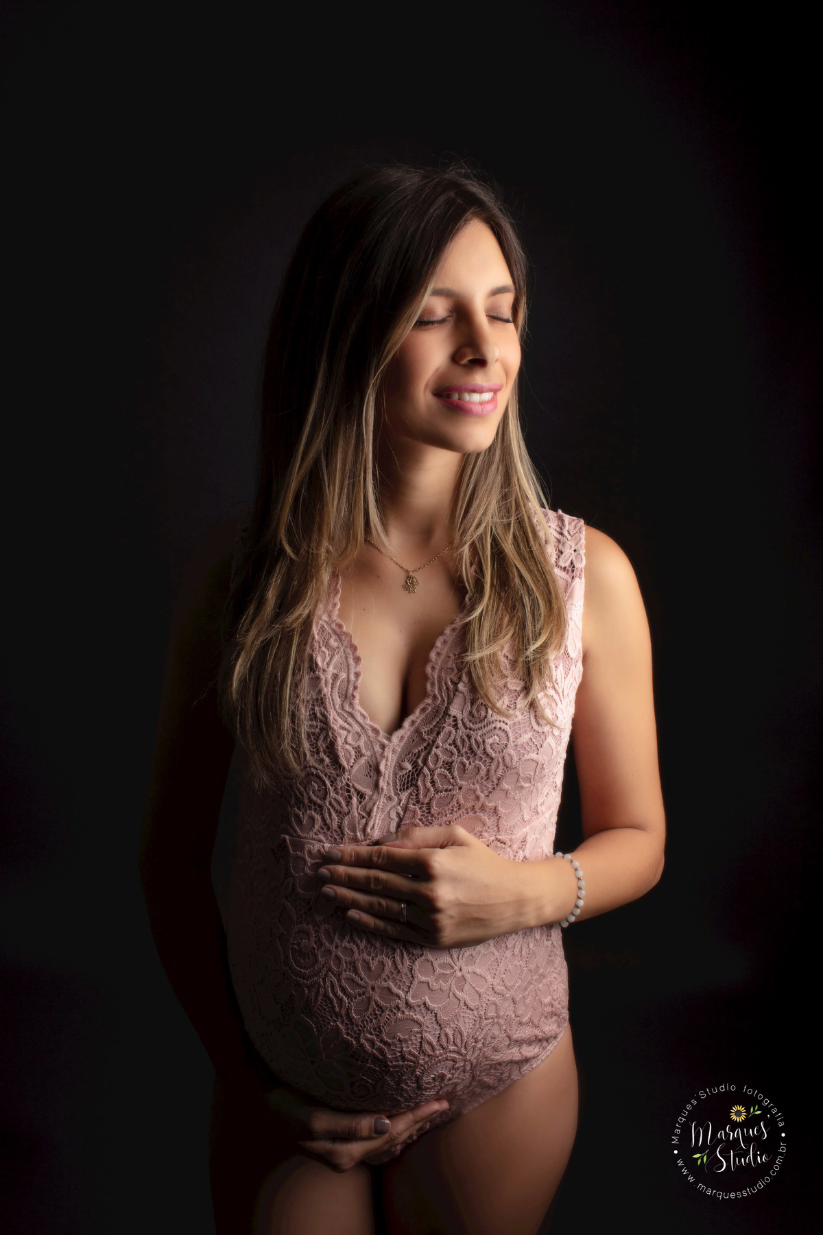 Foto feita em Studio na Vila Sumaré, São Paulo, SP, a gestante está em pé de perfil, ela está com as mãos sobre a sua barriga gravida com um belo sorriso estampado no rosto, ela está usando um body rose com renda, e está no fundo infinito preto.