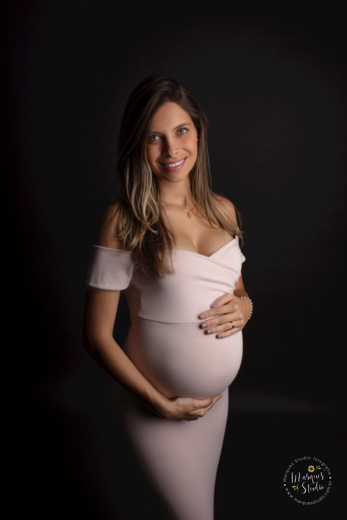 Foto feita no Studio em Pinheiros, São Paulo, SP, a gestante está em pé de perfil, ela está com as mãos sobre a sua barriga gravida, com um belo sorriso estampado no rosto, ela está usando um vestido longo rosa, e está no fundo infinito preto.