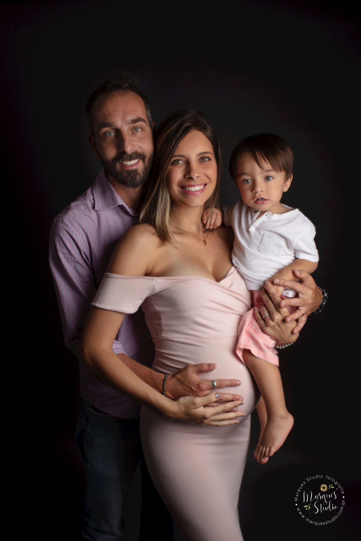 Fotografia de um casal de grávidos e seu bebê, a mamãe está na frente do papai, o pai está abraçando a mãe, a mamãe está com o irmão mais velho no colo, eles estão sorrindo, foto feita no studio localizado em Perdizes, São Paulo, SP.