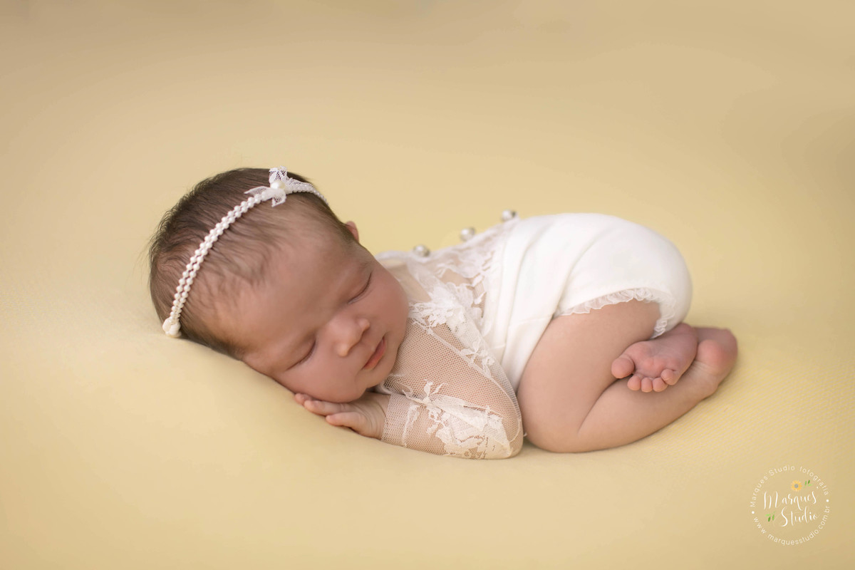 Fotografia de bebê recém-nascida menina, deitada no puff, com as mãozinhas no rosto, foto feita em studio fotográfico na Vila Perdizes, São Paulo, SP, ela está dormindo e usando uma linda roupinha branca com renda e um lindo lacinho com pérolas no cabelo.