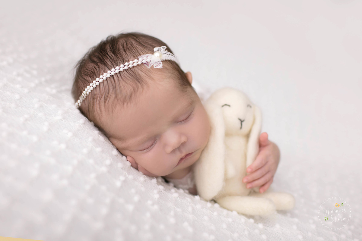 Fotografia de bebê menina, deitada no puff com uma manta branca de bolinhas, com as mão direita no rosto e a mão esquerda segurando um ursinho bege, foto feita em studio fotográfico localizado na zona Oeste, Osasco, São Paulo, SP, ela está dormindo.
