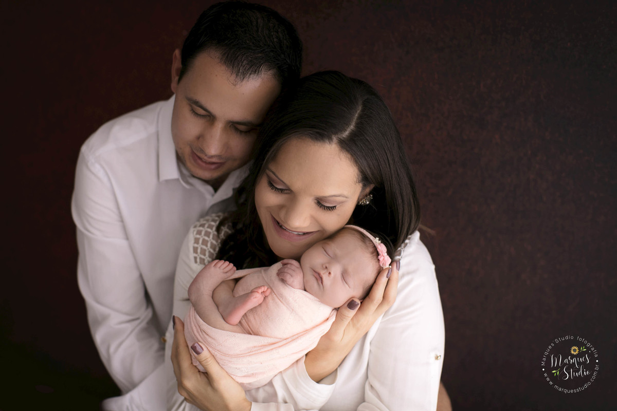 Fotografia de família no ensaio Newborn da Isadora de 18 dias, a mamãe está com a bebê no colo e o papai está  abraçando as duas, essa foto foi feita no fundo fotográfico da cor marrom, no estúdio localizado no Sumaré, São Paulo, SP.