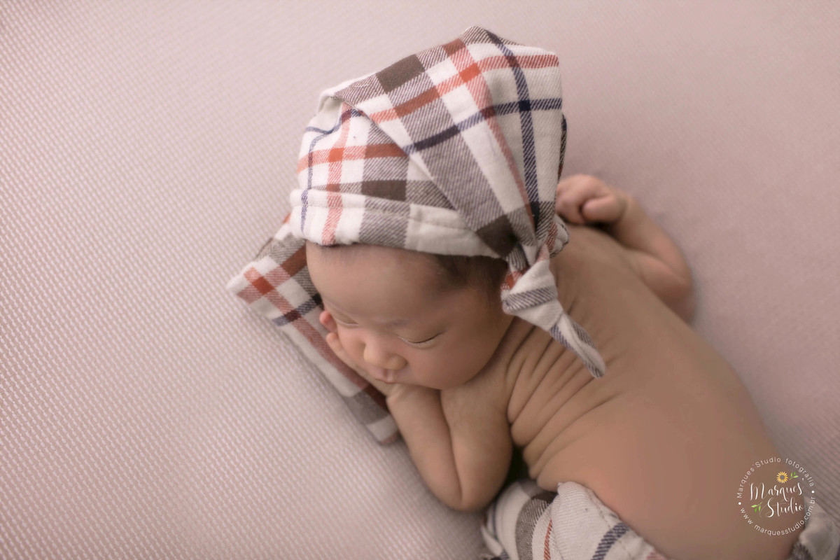 Fotografia de bebê recém-nascido menino, deitado no puff com as mãozinhas no rosto, foto feita em studio fotográfico na Pompéia, São Paulo, SP, ele está dormindo e usando uma linda calça com gorrinho xadrez, e com o rosto em um lindo travesseiro xadrez.