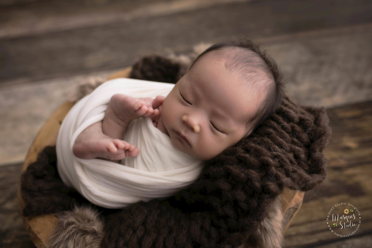 Fotografia feita para o ensaio Newborn do Thomas de 15 dias, feito no nosso studio localizado na Avenida Paulista, São Paulo, SP. Bebê no wrap bege, deitado em um cesto de madeira, ele está de barriga pra cima, no fundo fotográfico de madeira.