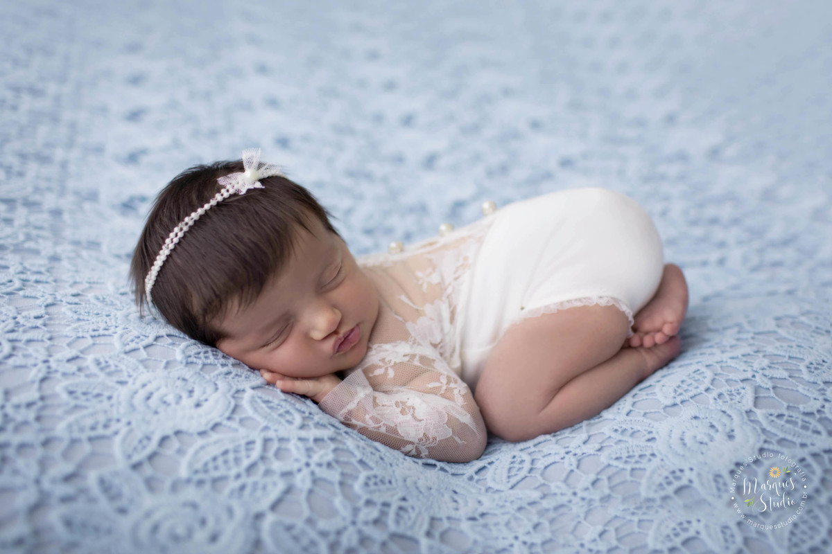 Fotografia de bebê recém-nascida menina, deitada no puff, com as mãozinhas no rosto, foto feita em studio fotográfico na Vila Perdizes, São Paulo, SP, ela está dormindo e usando uma linda roupinha branca com renda e um lindo lacinho com pérolas no cabelo.