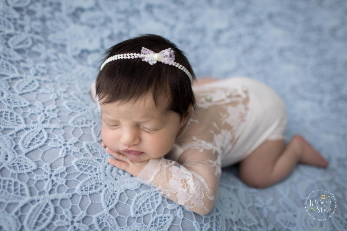 Foto de bebê menina, deitada no puff com uma manta de renda azul claro, com as mãos no rosto, foto feita em studio fotográfico localizado na Vila Leopoldina, São Paulo, SP, ela está dormindo e usando um lindo macacão na cor branca com renda.