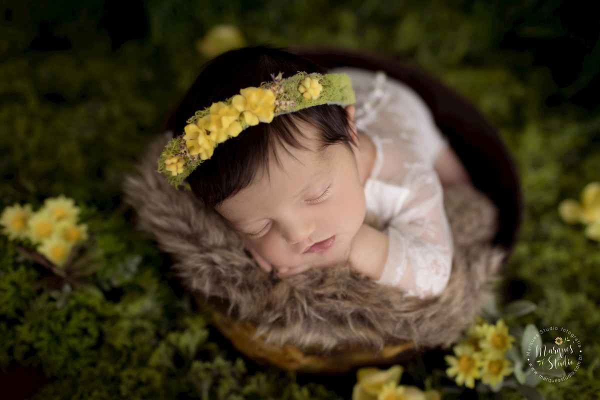foto de bebê recém-nascida no jardim, realizado no nosso studio fotográfico em São Paulo SP Vila Sumaré, a bebê está dormindo com as mãozinhas no rosto, em sua cabeça ela usa uma linda headband de florzinhas amarelas, e um lindo macacão branco de renda.