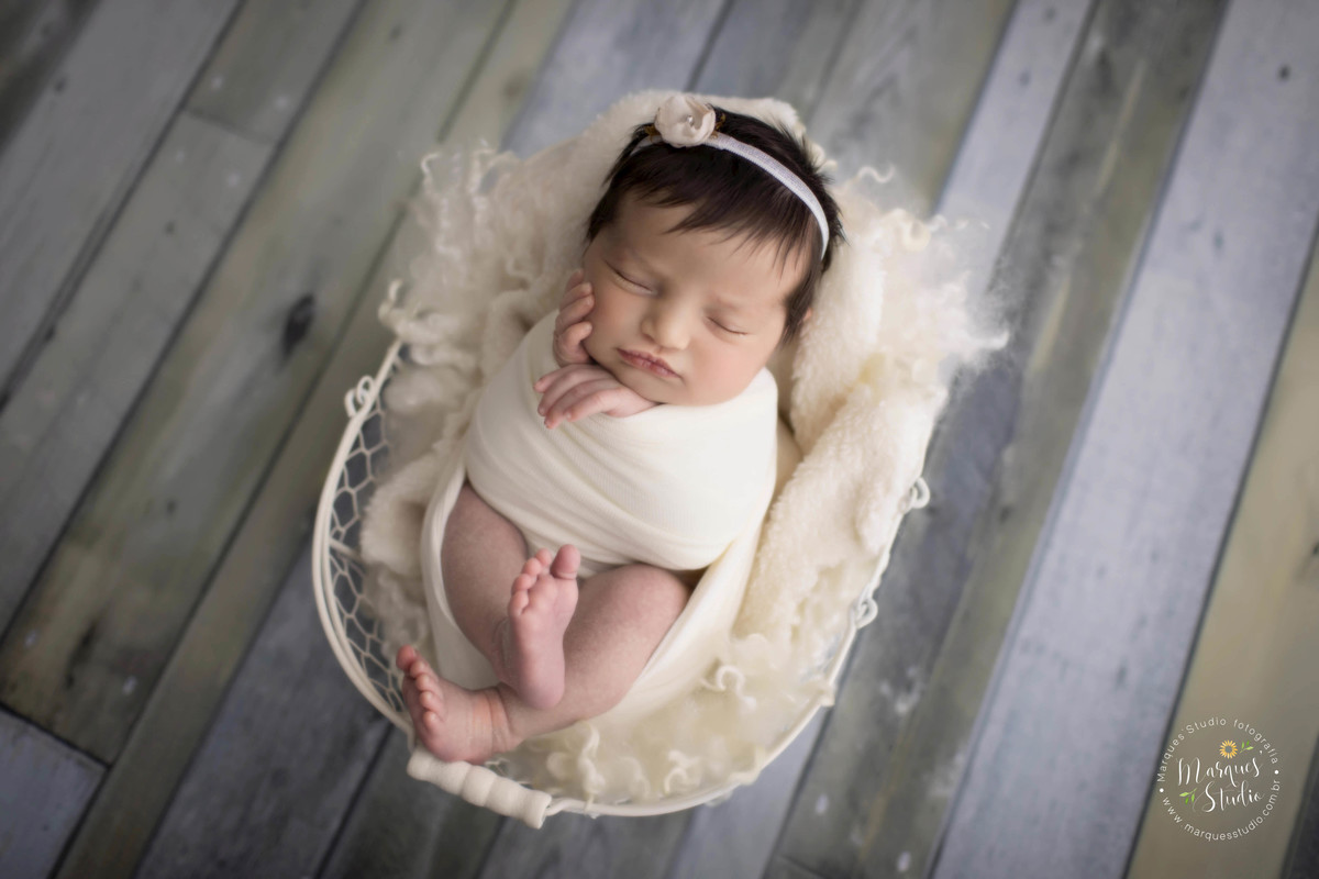 Ensaio Newborn feito no nosso studio em Pinheiros, São Paulo, SP. Bebê no wrap bege, ela está dormindo de barriga pra cima em um berço de cor branca. No lindo fundo fotográfico de madeira.