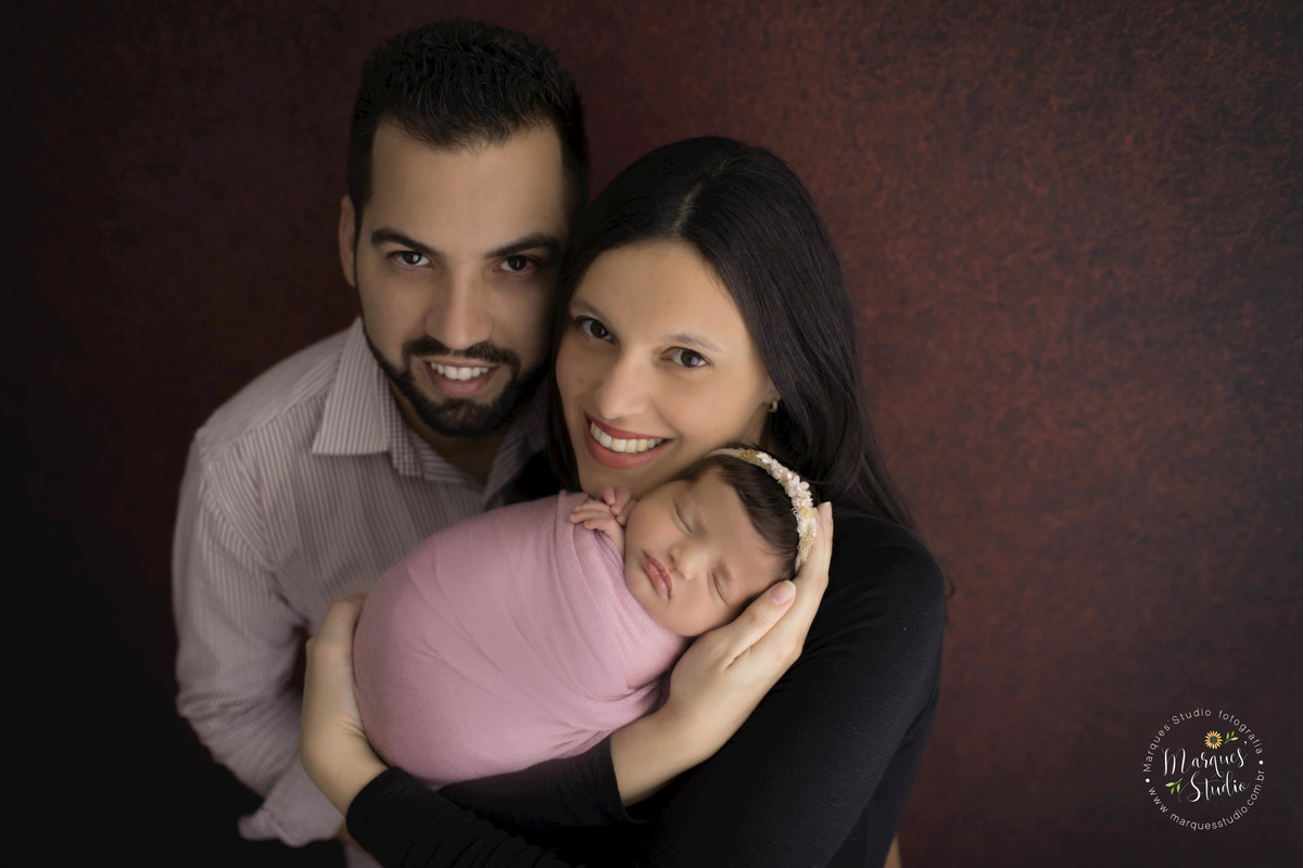 Fotografia de família no ensaio Newborn da Heloísa de 17 dias, a mamãe está com a bebê no colo e o papai está  abraçando as duas, essa foto foi feita no fundo fotográfico da cor marrom, no estúdio localizado na zona Oeste Osasco, São Paulo, SP.