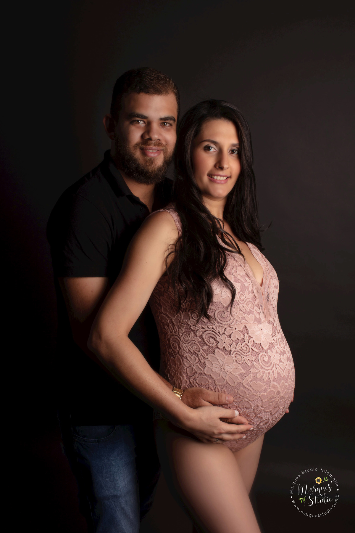 Fotografia de um casal de grávidos, a mamãe está na frente do papai, os dois estão com as mãos sobre a barriga da gestante, eles estão sorrindo, ela usa um body rose, e ele usa camiseta e calça jeans, eles estão no studio em Pinheiros São Paulo, SP.