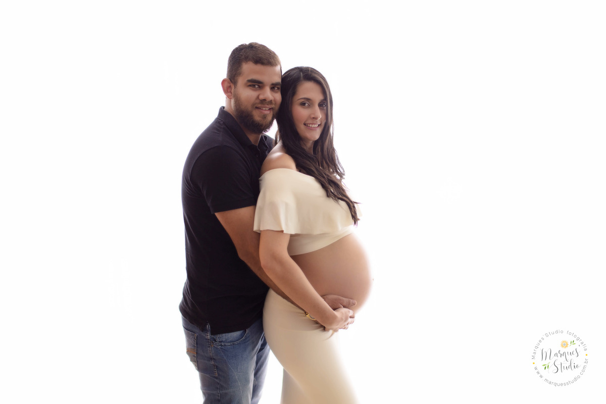Foto de um casal de grávidos, a mamãe está na frente do papai, os dois estão com as mãos sobre a barriga da gestante, eles estão sorrindo, ela usa um conjunto top e saia bege e ele usa camiseta e calça jeans, eles estão no studio no Morumbi São Paulo, SP.