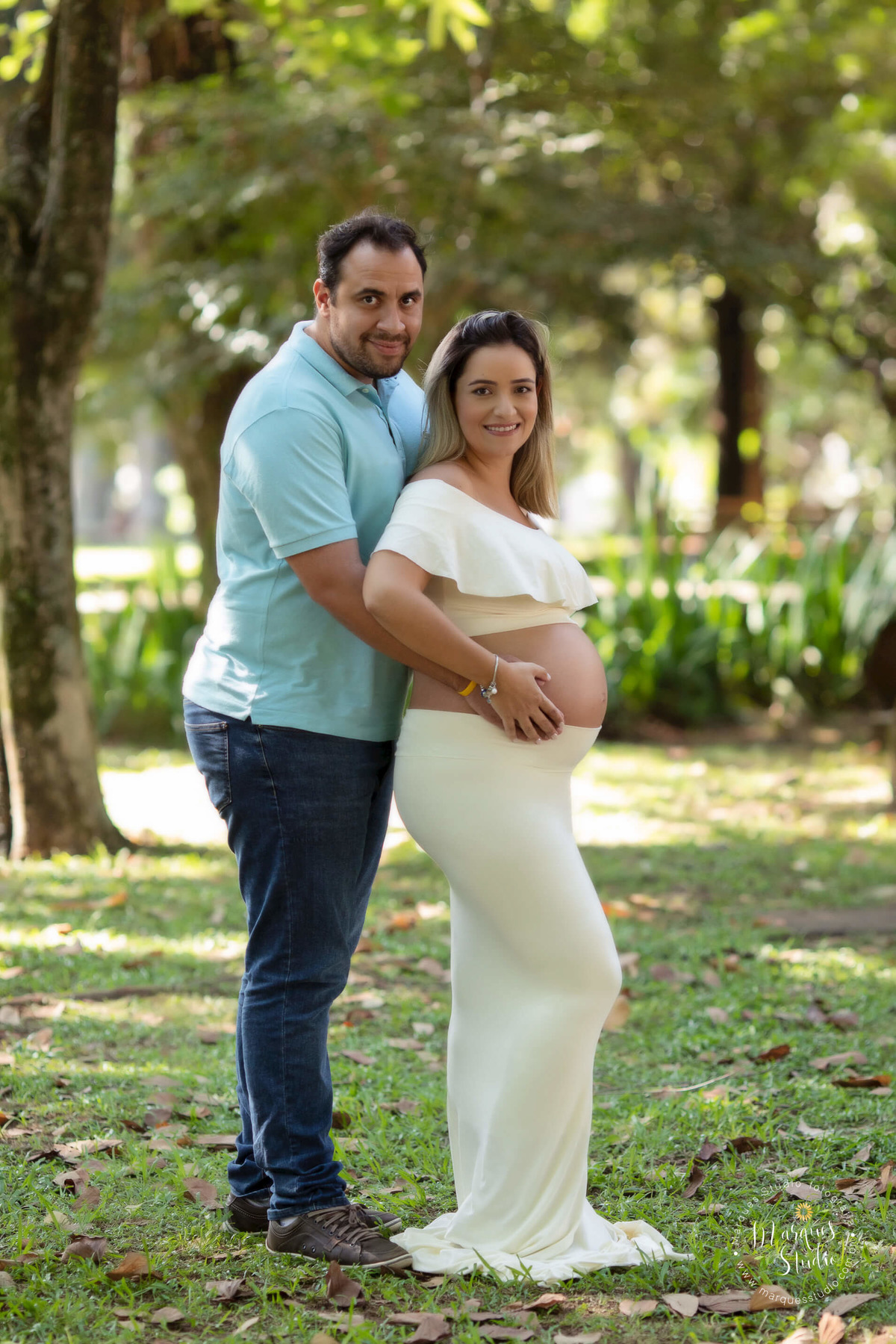 Foto feita no parque na Pompéia São Paulo SP com um casal de grávidos eles estão abraçados ela está na sua frente e ambos estão com as mãos sobre a barriga da gestante eles estão sorrindo, ela usa um conjunto de top e saia longa na cor creme.