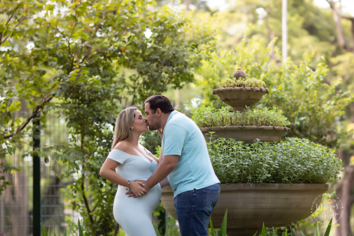 Fotografia feita na sessão externa no Morumbi São Paulo SP com um lindo casal de grávidos, eles estão um de frente para o outro com as mãos sobre a barriga da gestante e estão se beijando, ela usa um lindo vestido longo azul, e ele calça jeans e camisa.
