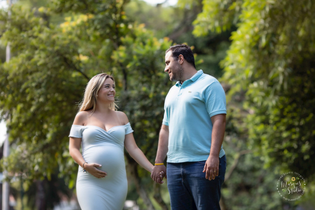 Foto de um lindo casal de grávidos no parque, eles estão de mãos dadas, se olhando e sorrindo, ela está usando um lindo vestido longo azul claro, do nosso studio na Vila Leopoldina, SP, temos vários modelos e cores de vestidos disponíveis para os ensaios.