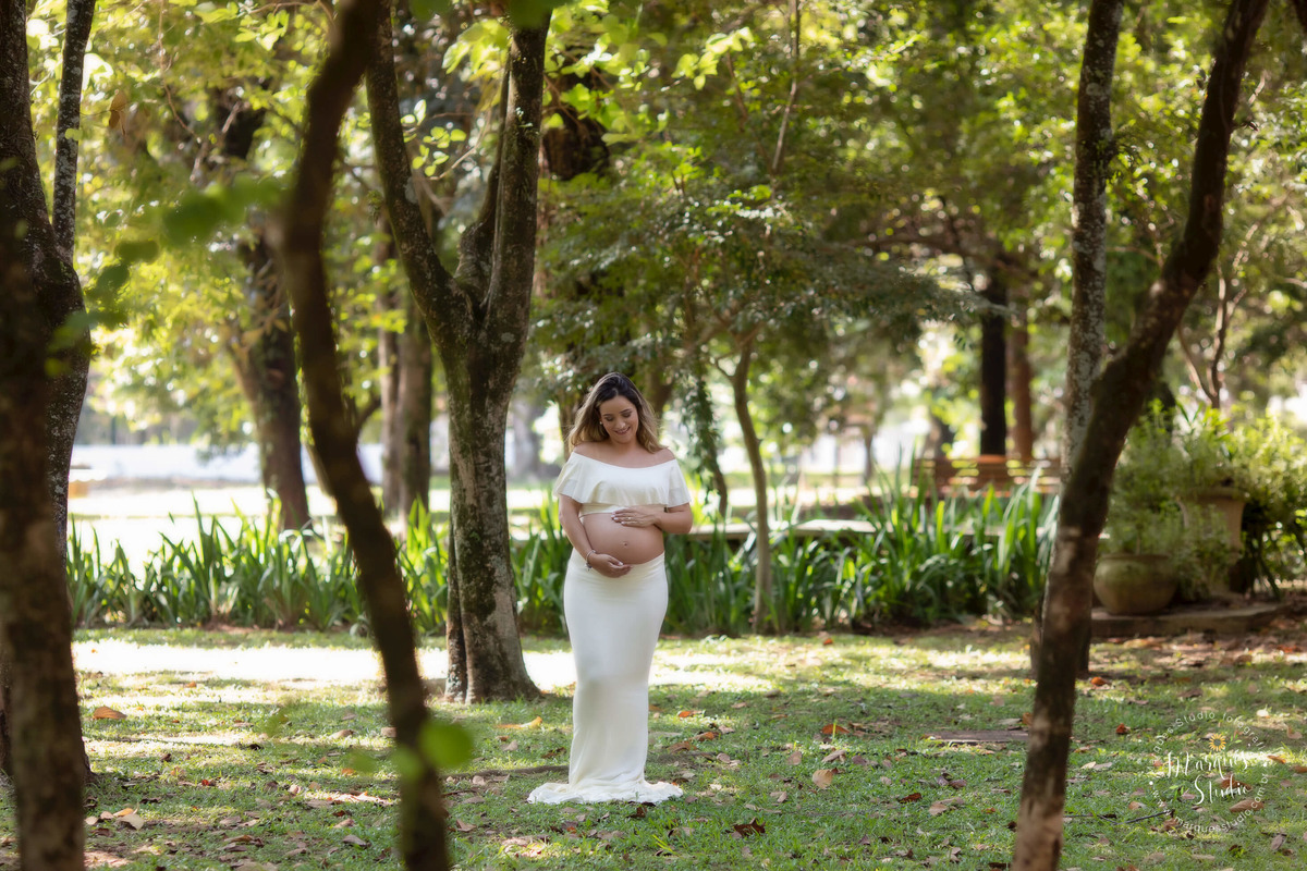 Fotografia de gestante no parque de São Paulo SP Vila Sumaré o ensaio externo foi feito no parque com muitas arvores e folhas ao fundo, a gestante está usando um belíssimo conjunto de top e saia longa na cor bege, ela está com as mãos sobre a sua barriga 