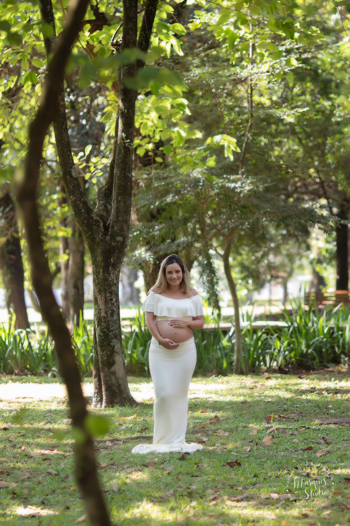 Fotografia de gestante no parque de São Paulo SP Av. Paulista a sessão externa foi feita no parque com muitas arvores e folhas ao fundo, a gestante está usando um belíssimo conjunto de top e saia longa na cor bege, ela está com as mãos sobre a sua barriga