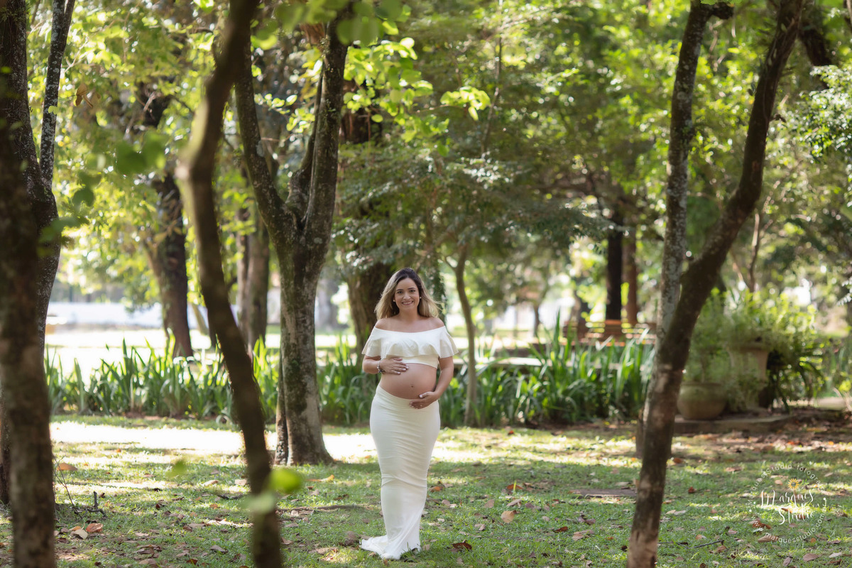 Foto feita na região Oeste de São Paulo SP Vila Madalena para o ensaio gestante da Cristina, ela está usando um belo conjunto de top e saia na cor bege com as mãos sobre a sua barriga dando um lindo sorriso, ao fundo tem lindas arvores com folhas  verdes.