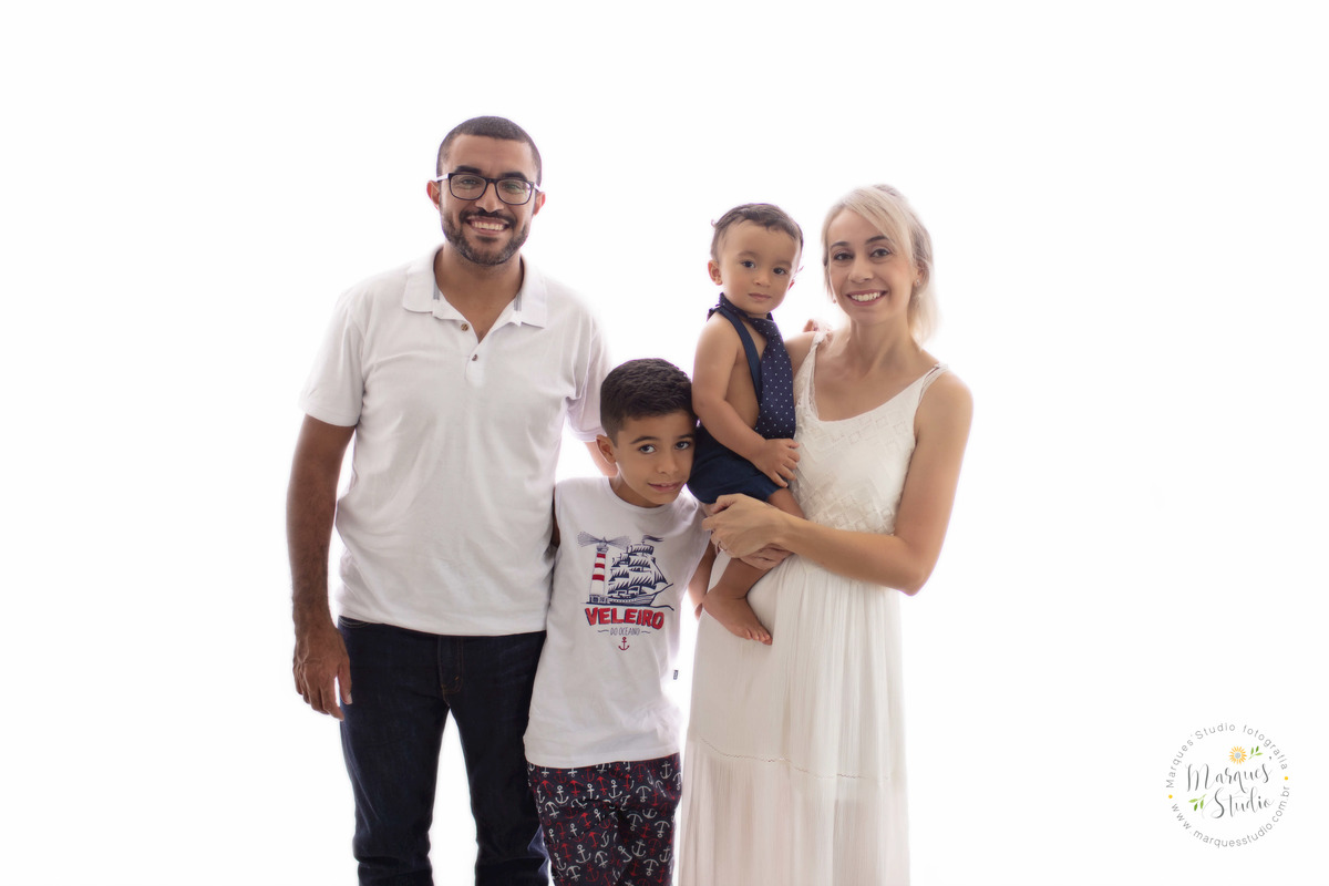 Fotografia da sessão de acompanhamento do bebê Theo de 12 meses, ele está com a mamãe o papai e o irmão mais velho em um fundo fotográfico branco, a família está abraçada e sorrindo, eles estão no studio localizado na Vila Leopoldina, São Paulo, SP.