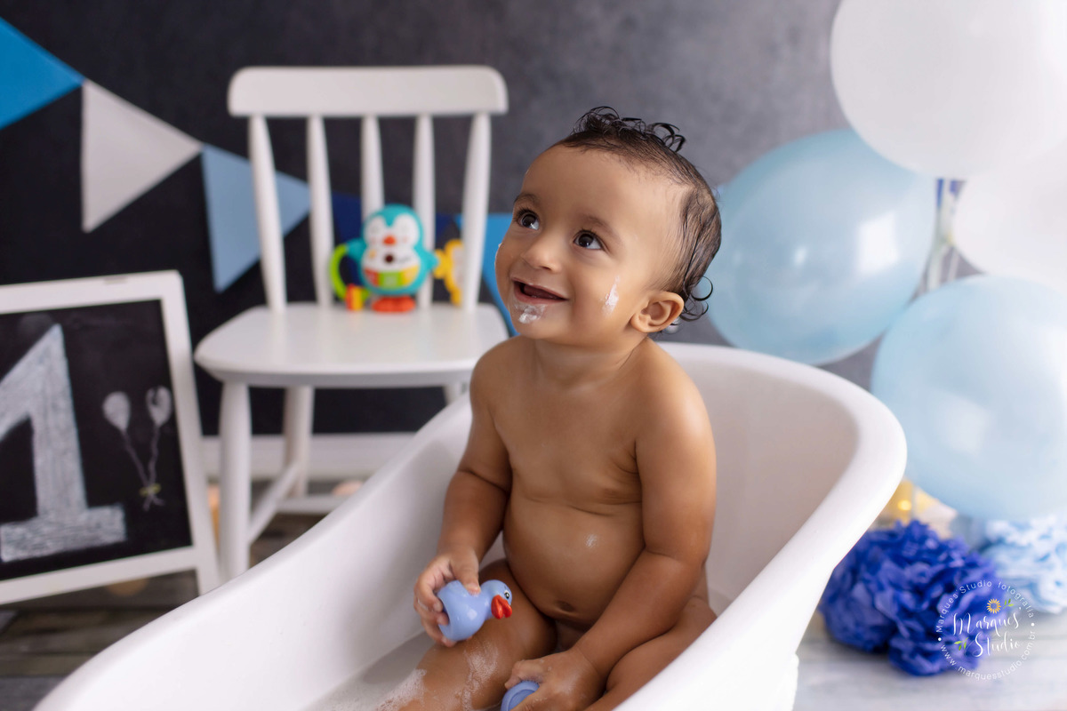 Foto do bebê Theo de 12 meses na sua sessão de acompanhamento, ele está sentado em uma linda banheira branca, tomando banho e está sorrindo, ao fundo tem uma cadeirinha branca uma lousa, bandeiras e balões azul e branco, o studio fica na Pompéia São Paulo