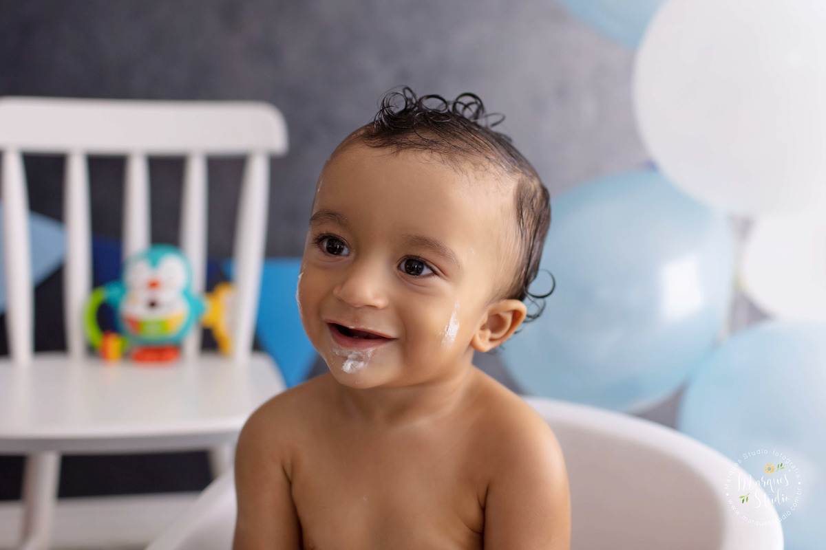 Foto Feita para a sessão Smash The Cake no studio, na zona Oeste, Osasco, SP, de uma criança de 1 ano ele está sentado em uma banheira branca, e está olhando para o lado direito dele, com balões e bandeirinhas ao fundo nas cores azul e branco, ele sorriu.