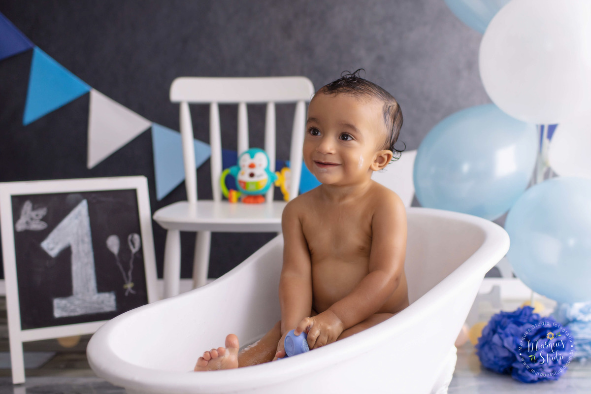 Foto do bebê Theo de 1 aninho na sua sessão de acompanhamento, ele está sentado em uma linda banheira branca, tomando banho e está sorrindo, ao fundo tem uma cadeirinha branca uma lousa, bandeiras e balões azul e branco o studio fica em Perdizes São Paulo