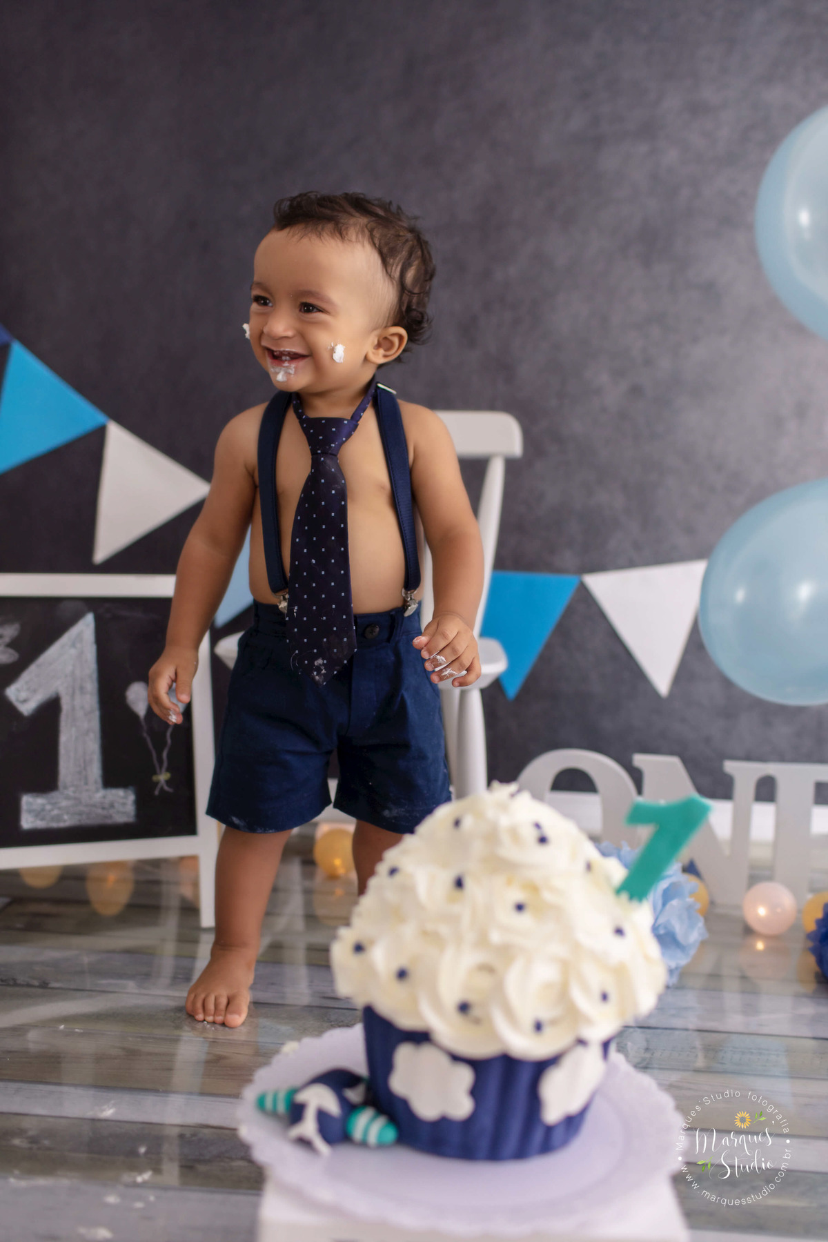 Fotografia do Bebê Theo de 1 aninho no ensaio Smash The Cake no studio localizado São Paulo SP Capital, ele está atras do bolo nas cores azul e branco com um aviãozinho decorativo, ao fundo uma cadeira branca balões e bandeiras azuis e branco, ele sorri.