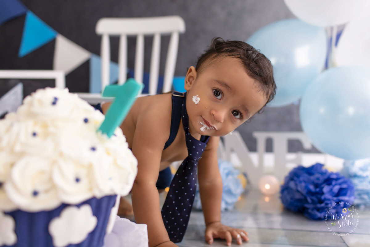 Foto do Bebê Theo de 12 meses no ensaio Smash The Cake no studio localizado na Av. Paulista SP ele está atras do bolo nas cores azul e branco com nuvens decorativas, ao fundo uma cadeira branca balões e bandeiras azuis e branco, ele está com bolo na boca