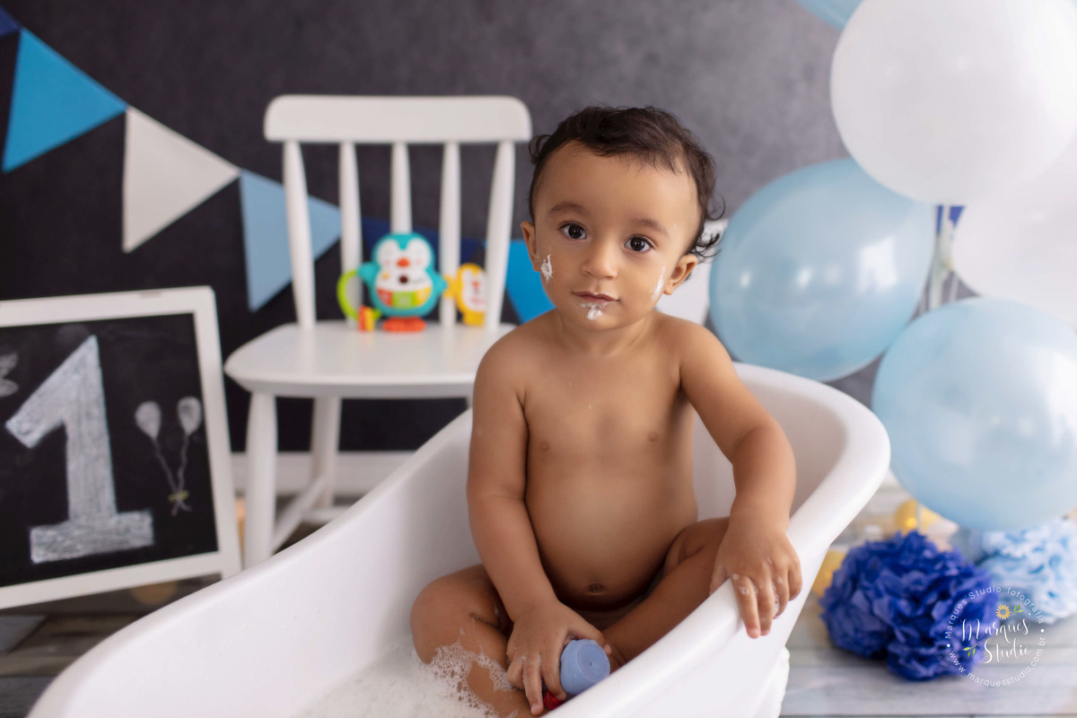 Fotografia do Theo de 12 meses na sua sessão de acompanhamento, ele está sentado em uma linda banheira branca, tomando banho, ao fundo tem uma cadeirinha branca uma lousa, bandeiras e balões azuis e branco, o studio fica na Vila Leopoldina, SP.