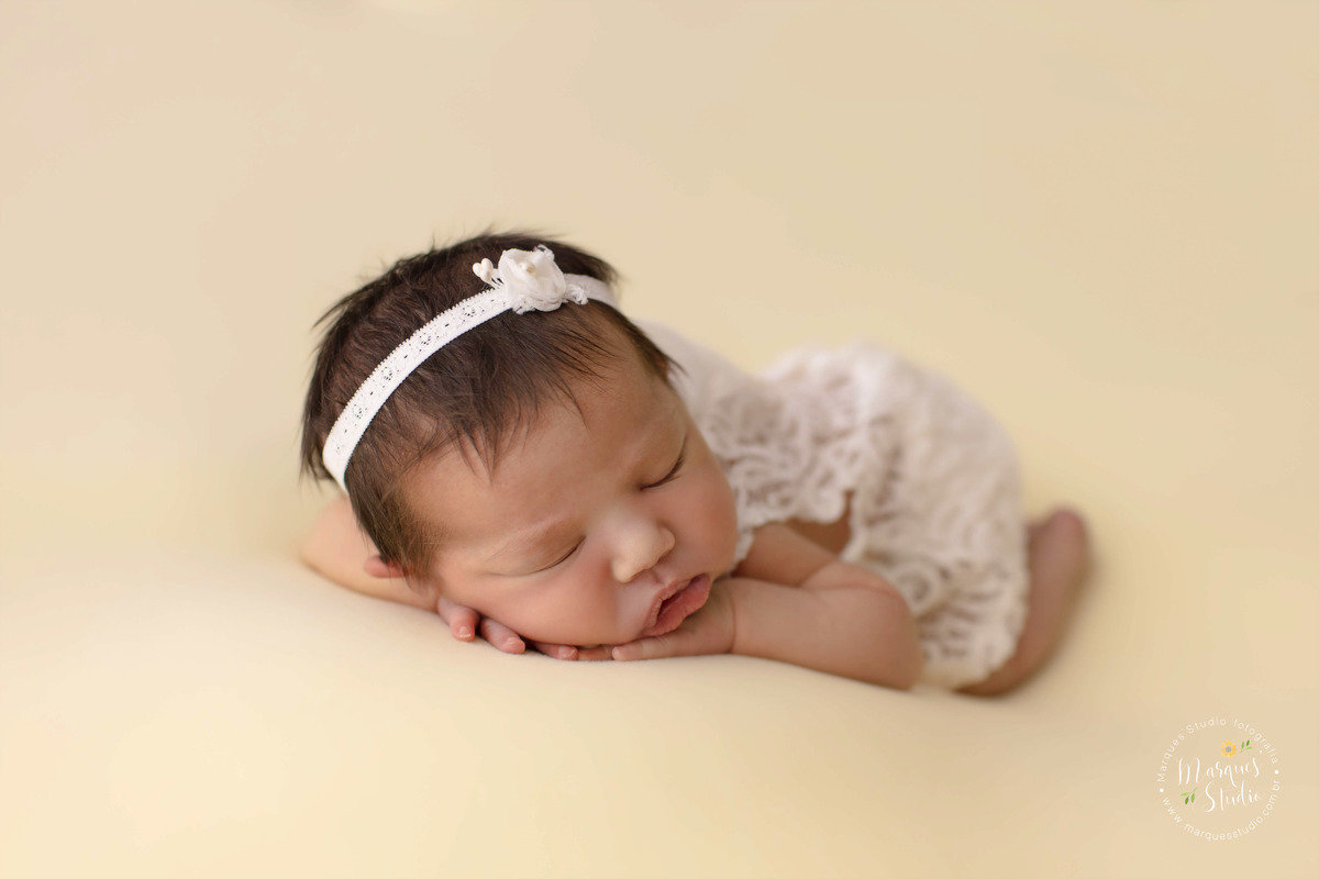 Fotografia de bebê menina, deitada no puff com as mãos no rosto, foto feita em studio fotográfico na Avenida Paulista, São Paulo, SP, ela está dormindo e usando um lindo vestido na cor branca com renda, e no cabelo uma linda headband de lacinho branco.