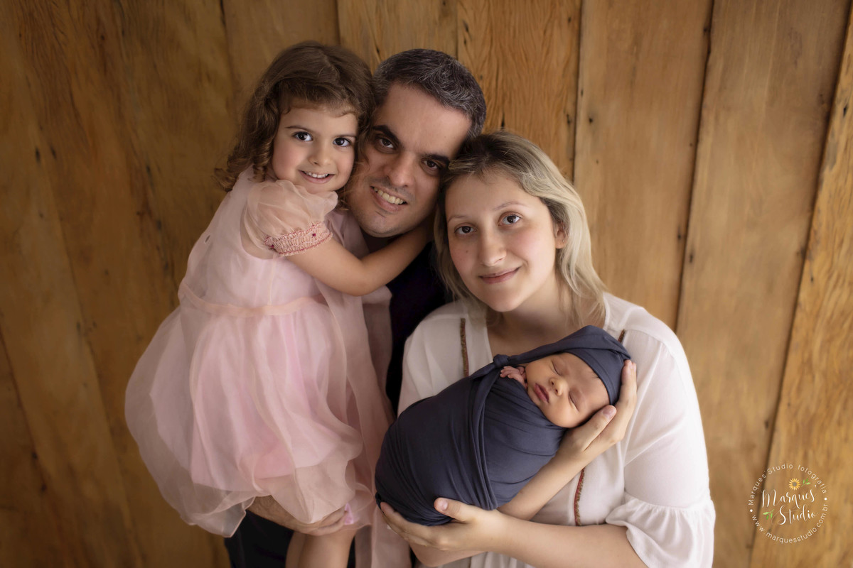 Fotografia de família no ensaio Newborn, a mamãe está com o bebê no colo e o papai está com a irmã no colo, eles estão abraçados, eles estão sorrindo, essa foto foi feita no fundo fotográfico de madeira, no estúdio localizado no Sumaré, São Paulo, SP 