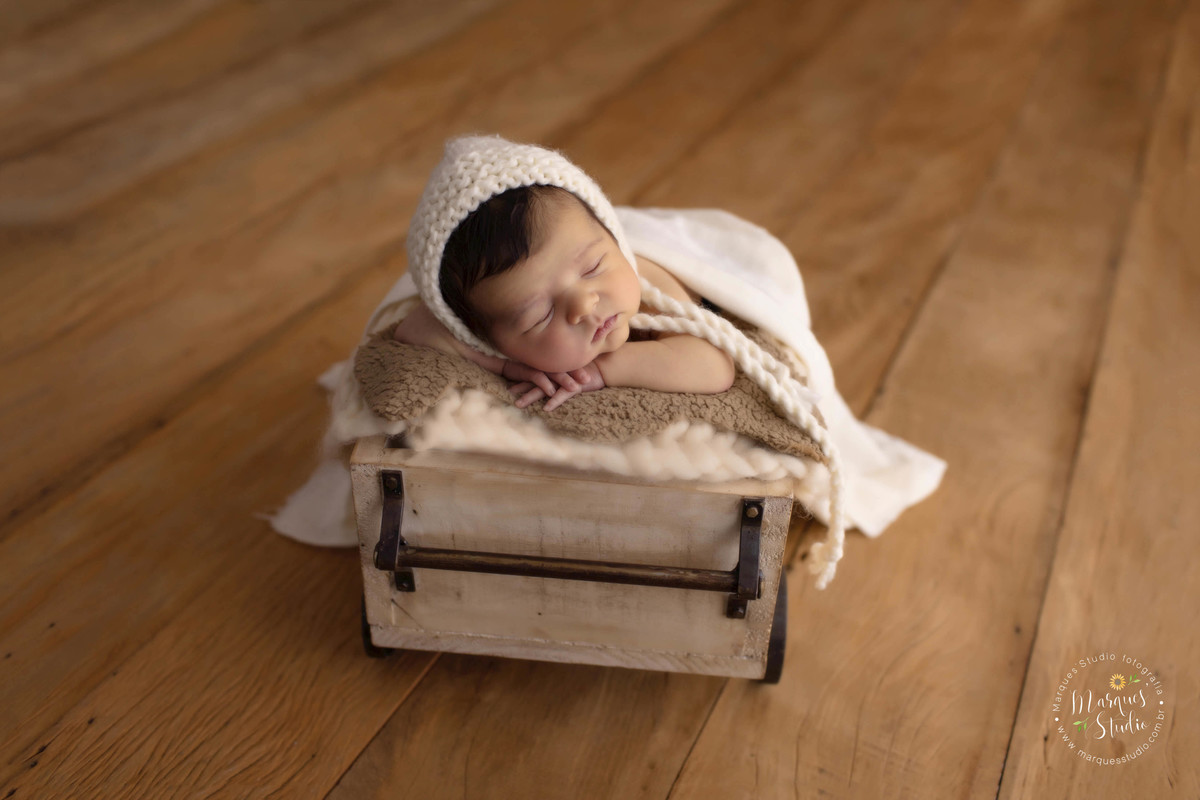 Fotografia do bebê deitado em vagãozinho, em um fundo fotográfico de madeira. Foto feita em Estúdio na zona Sul de São Paulo - SP, ele está dormindo de barriga pra baixo, com as mãozinhas no rosto, ele usa uma linda touca bege e está coberto com uma manta