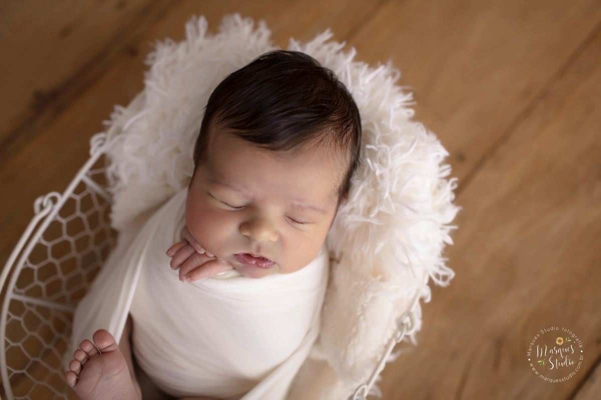 Ensaio newborn feito no nosso studio na zona Oeste de São Paulo, Vila Leopoldina SP. Bebê no wrap de cor bege, de barriga pra cima, ele está deitado e dormindo em um lindo cesto na cor branca, em um fundo fotográfico de madeira.