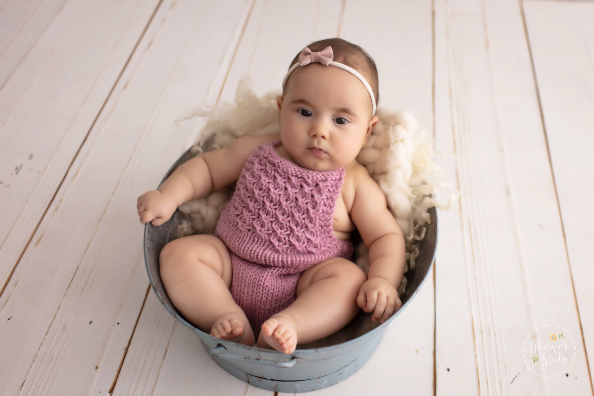 Foto feita em studio fotográfico localizado na zona Oeste, Osasco, São Paulo, SP, com uma criança de 4 meses sentada em um cesto azul, ela está sorrindo, o fundo fotográfico é de madeira, ela usa um lindo macacão rose de crochê e lacinho rosa na cabeça.
