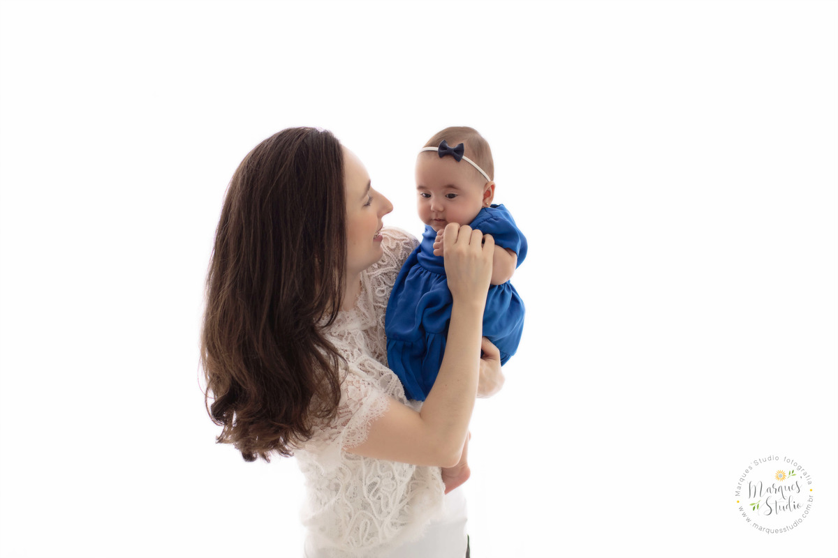 Foto da sessão de acompanhamento de 4 meses da Bia, no Studio na Vila Madalena, São Paulo, SP, na foto temos a mamãe de perfil segurando a bebê no colo que está dando um lindo sorriso, elas estão no fundo fotográfico infinito, a bebê usa um vestido azul.
