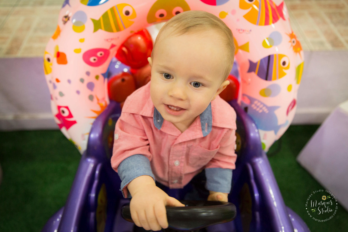 Foto feita no aniversário do Benjamin de 1 ano. No salão de festas que fica localizado na Vila Leopoldina - São Paulo, SP. Na foto o aniversariante está brincando sorridente, em um carrinho da cor roxa.