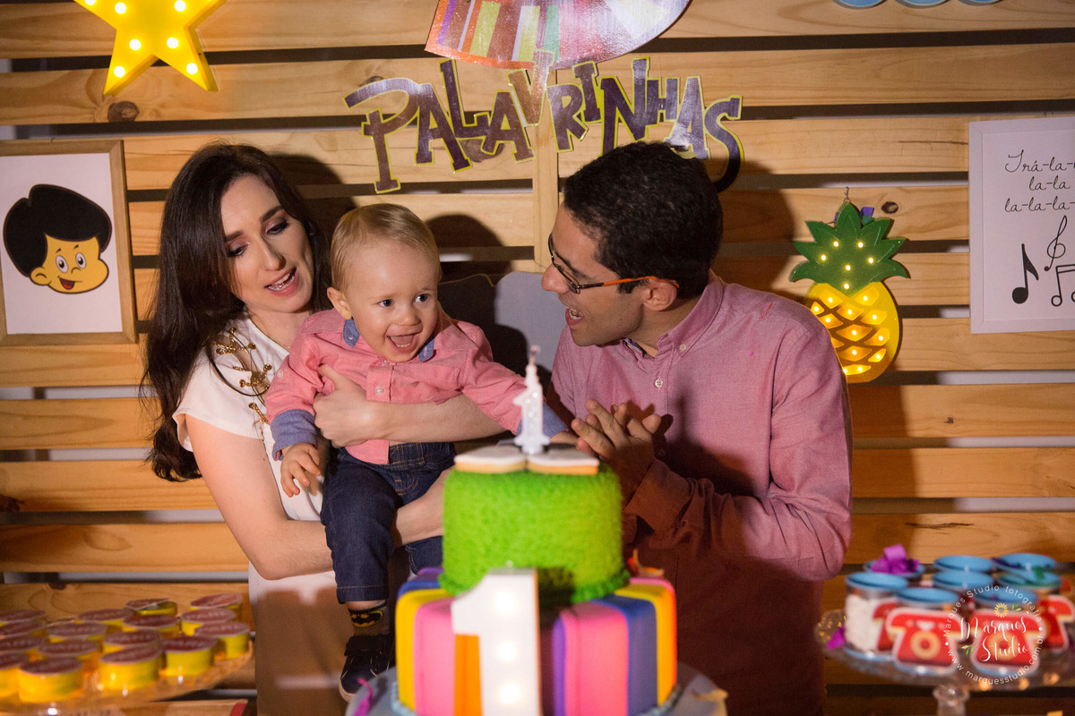 Fotografia feita no aniversário do Benjamin de 1 ano. Na foto o aniversariante está com seus pais cantando parabéns. Eles estão atrás da mesa do bolo decorada com o tema 3 palavrinhas. O buffet está localizado na Consolação - São Paulo, SP