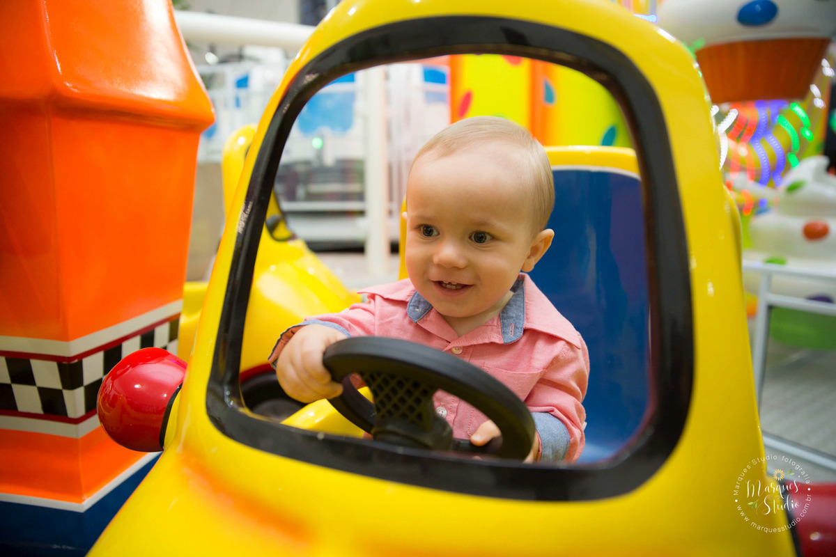 Foto feita no aniversário do Benjamin de 1 ano. No salão de festas que fica localizado na Pompeia - São Paulo, SP. Na foto o aniversariante está brincando sorridente, em um carrinho da cor amarela.