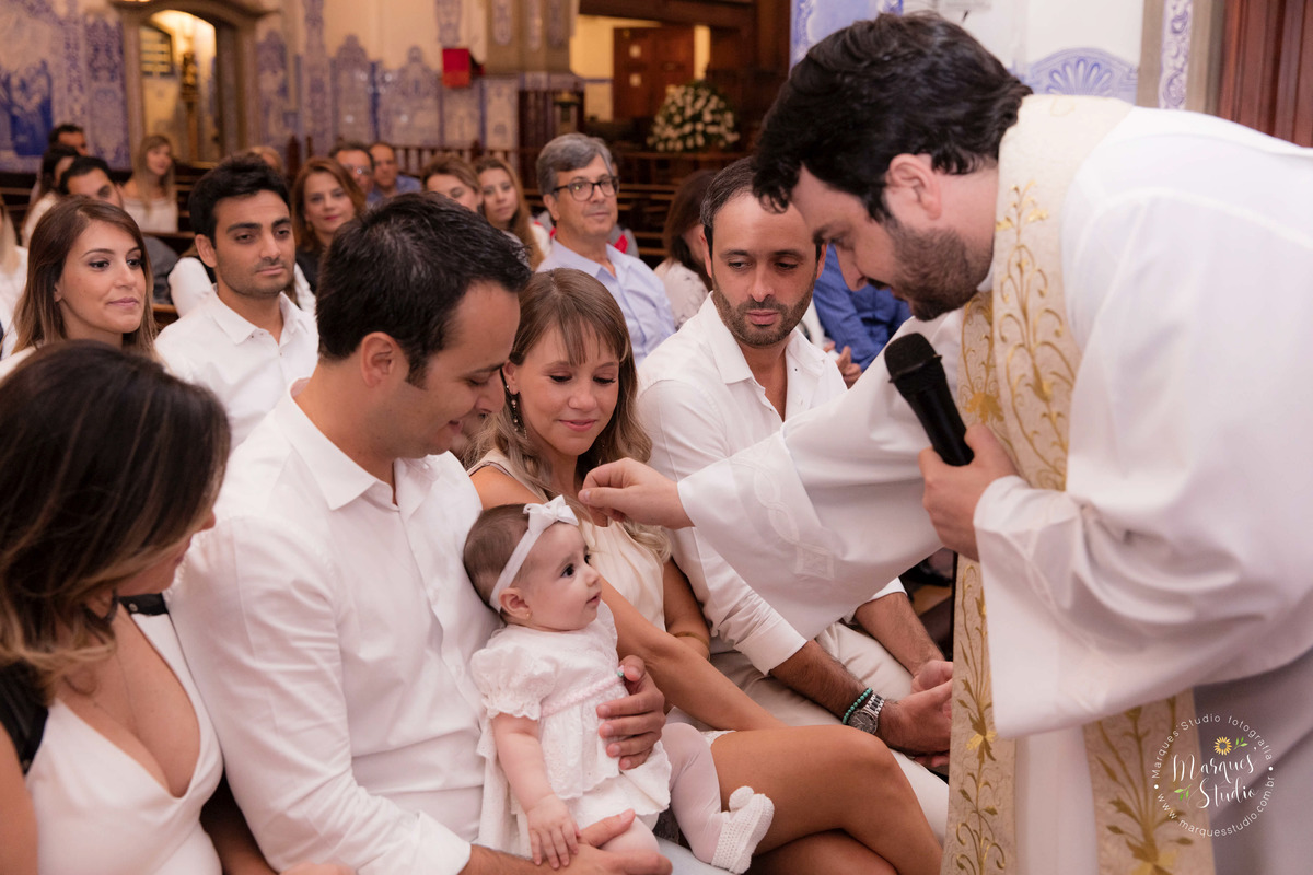 Foto tirada no batizado da Pietra na igreja Nossa Senhora do Brasil em São Paulo, SP. Na foto temos os pais, os padrinho, o padre e a Pietra. O padre está fazendo o sinal da cruz na bebê. 