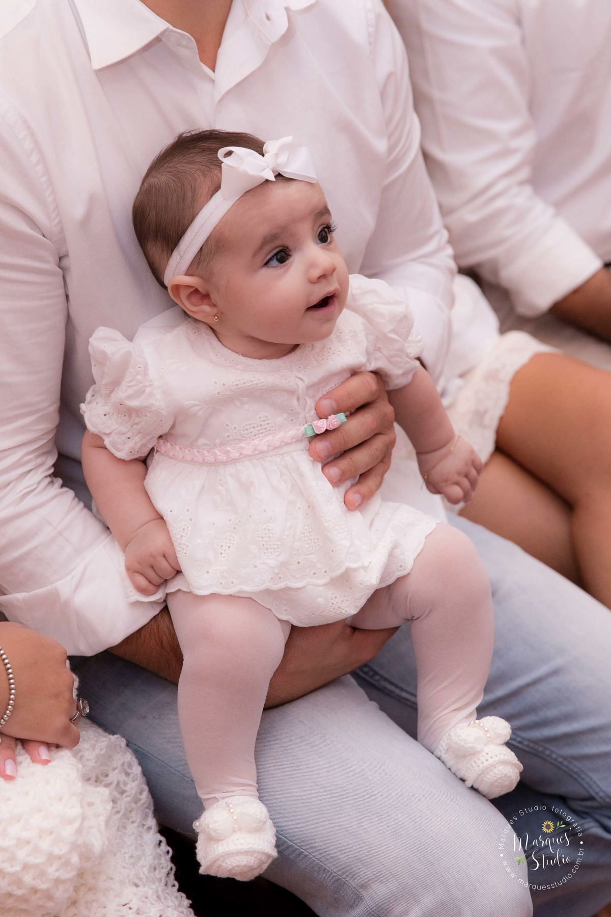 Fotografia feita no batizado da Pietra. Na foto temos a Pietra no seu lindo vestido branco com detalhes rosa, com um delicado laço branco na cabeça e dando um sorriso. A igreja está localizada na Av Paulista em São Paulo, SP.