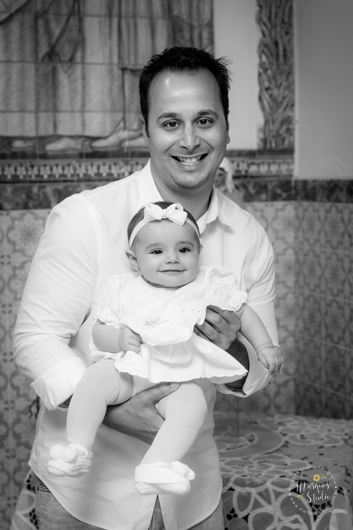 Fotografia feita no batizado da Pietra, na igreja localizada na Vila Madalena, em São Paulo, SP. Na foto temos a Pietra no colo do seu papai, dando um lindo sorriso. Ele estão muito felizes. 