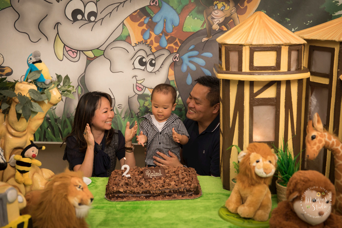 Foto registrada no aniversário de 2 anos do Rafael, em um buffet localizado na Vila Leopoldina - São Paulo, SP. Na foto temos a família com o aniversariante atrás da mesa do bolo cantando parabéns. O tema da festa foi safari.