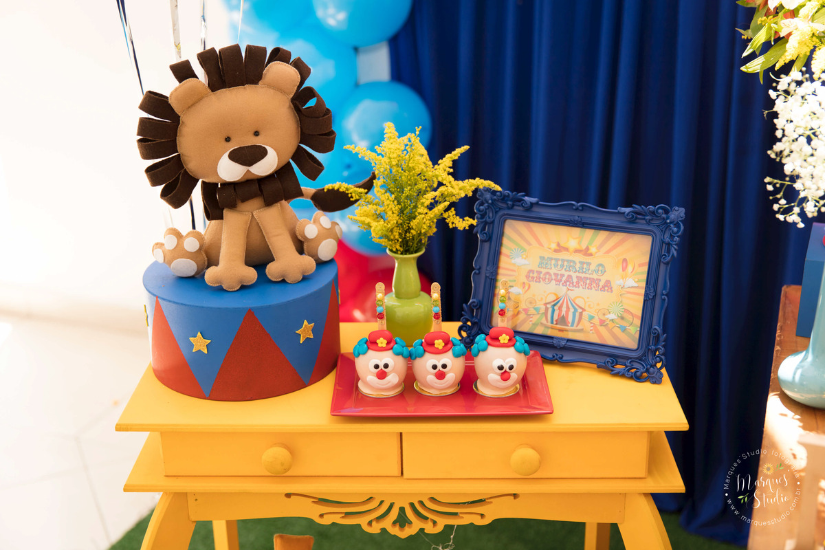 Fotografia tirada no aniversário dos gêmeos Murilo e Giovanna na Zona Sul de São Paulo, SP. Na foto temos parte da decoração com o tema circo, com balões coloridos, animais de pelúcia, docinhos e um porta retrato com nome dos aniversariantes.  