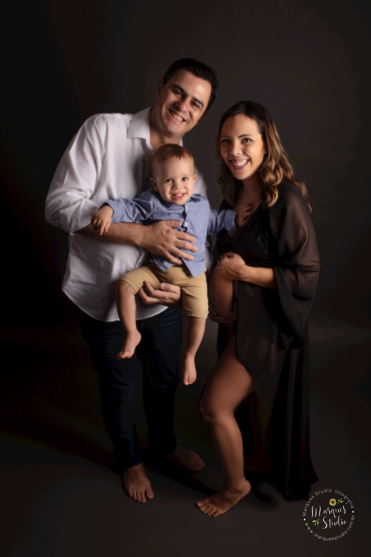 Fotografia de um casal de grávidos e seu bebê, a mamãe está com as mãos na barriga, o papai está segurando o irmãozinho mais velho no colo, eles estão sorrindo. Foto feita no studio localizado em Perdizes, São Paulo, SP.