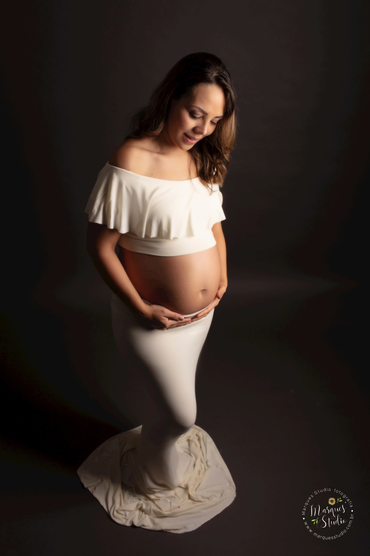 Foto feita para o ensaio da gestante Erika no studio de fotografia em Osasco São Paulo, SP, Ela está usando um lindo conjunto de saia e blusa bege. Ela está em pé com as mãos sobre a sua barriga de gestante, e está dando um lindo sorriso para o seu bebê.
