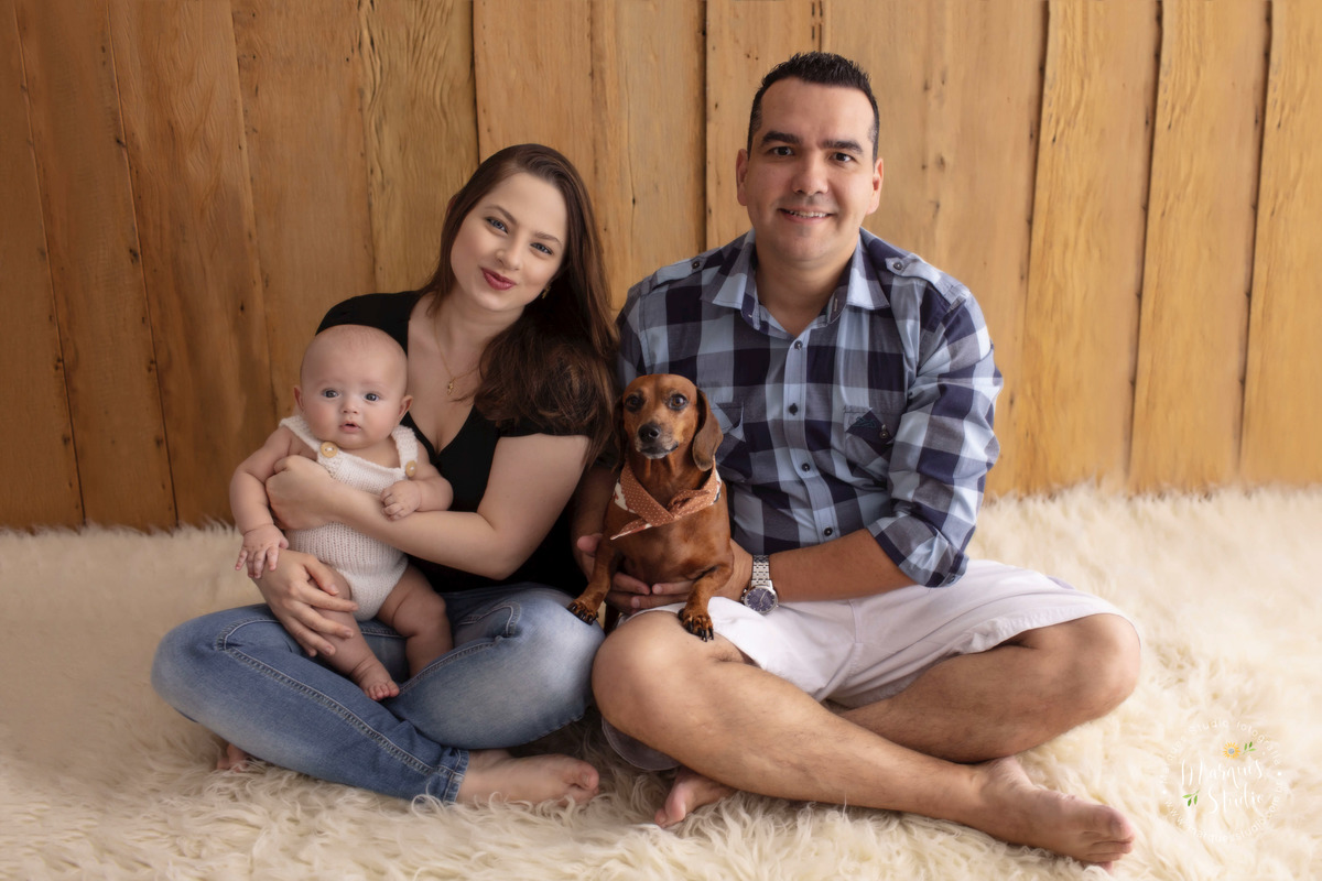 Foto tirada no ensaio do Enrico de 03 meses. Na foto temos toda a família do Enrico abraçadinha. A mamãe está segurando o bebê no colo e o papai a cachorrinha. Eles estão sentados em um tapete branco. Studio encontra-se na Zona Sul de São Paulo, SP