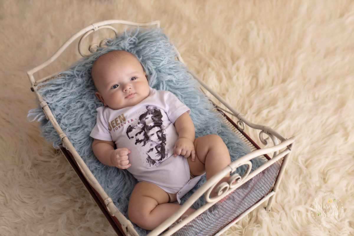 Fotografia feita no ensaio de bebê do Enrico. Na foto ele está deitado em um berço com uma manta azul, em cima de um tapete branco. Ele está vestido com um macacão da banda Quens. Studio localizado na Av. Paulista - São Paulo, SP.