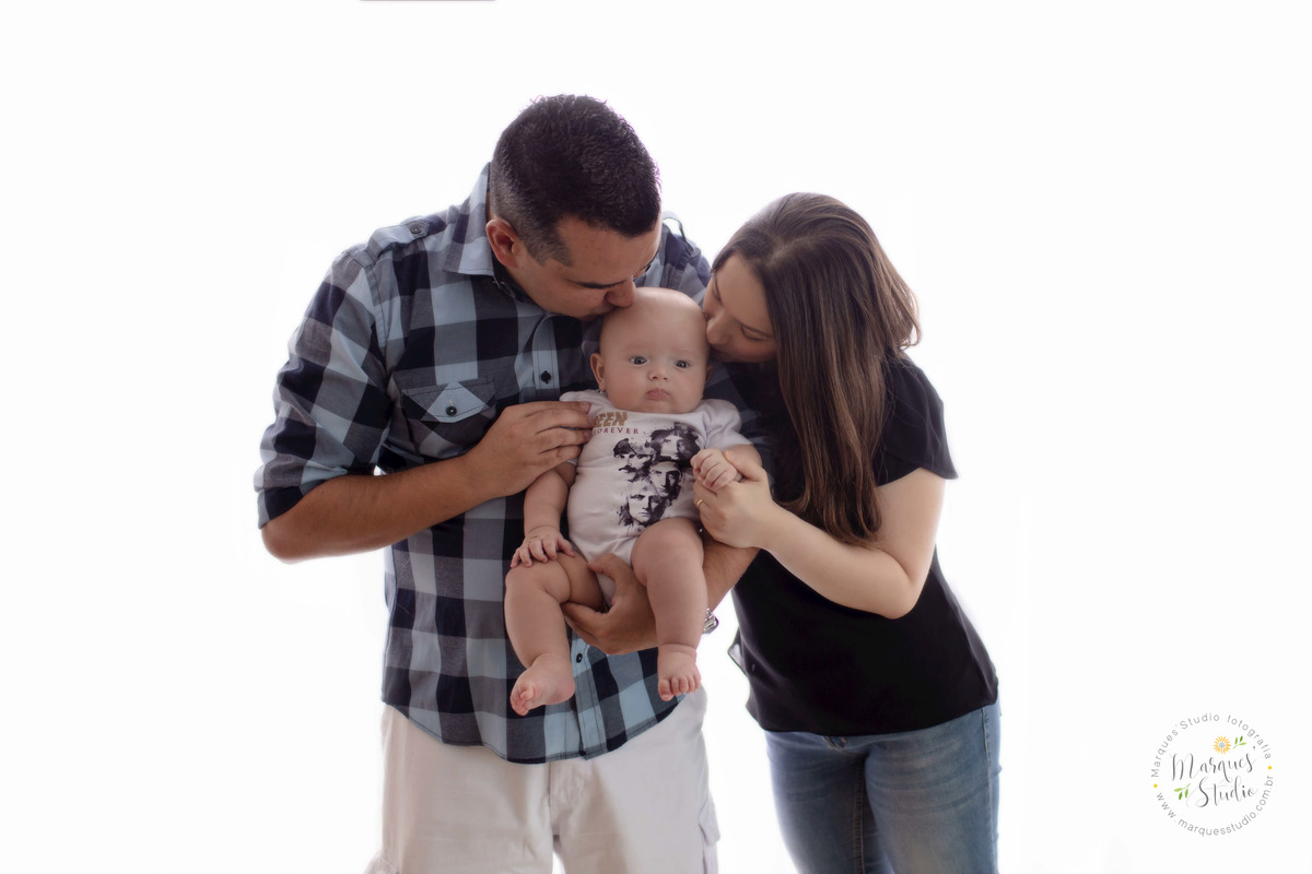 Foto tirada no studio que está localizado na Pompeia - São Paulo, SP. Na foto o papai está segurando o bebê no colo e ele e a mamãe dão um beijinho na cabeça do bebê.
