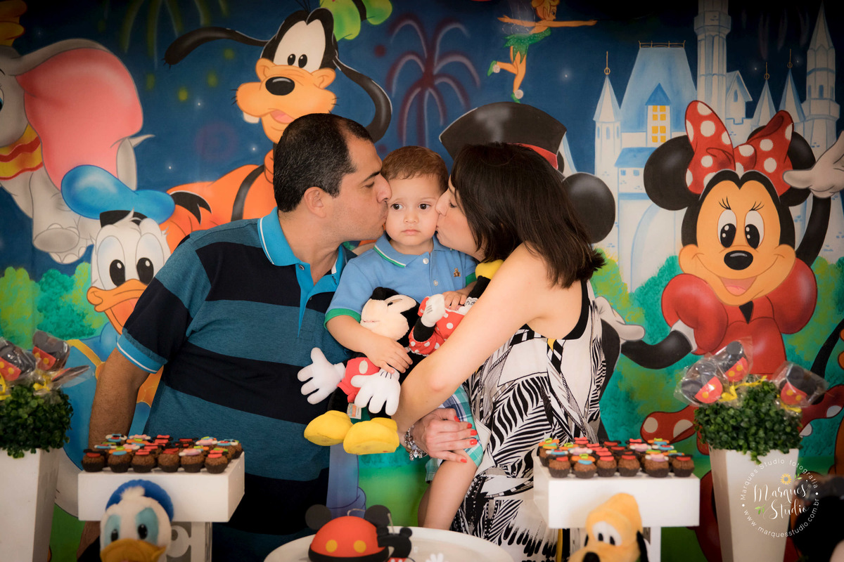 Fotografia registrada no aniversário do Guilherme de dois aninhos. Na foto temos o Guilherme no colo da mamãe, recendo um beijão da mamãe e do papai. Eles estão atrás da mesa do bolo em buffet que está localizado em Santo Amaro - São Paulo, SP