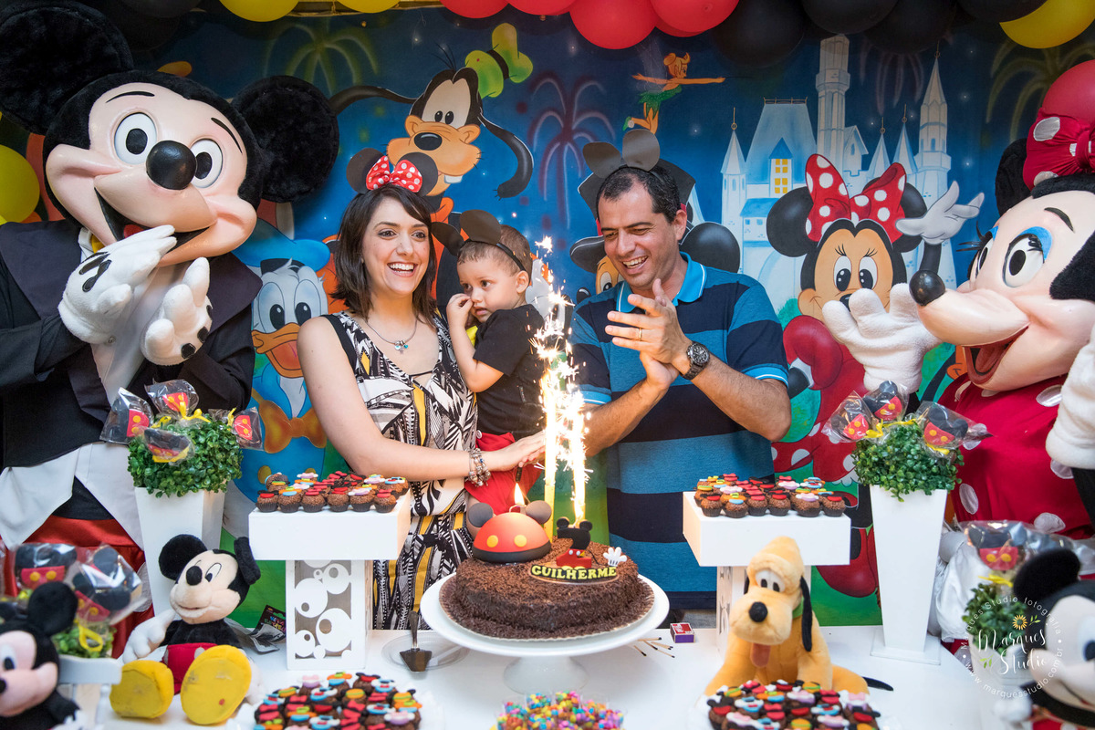 Fotografia tirada no aniversário de 2 aninhos do Guilherme em um Buffeet localizado em Santo Amaro - São Paulo, SP. Na foto temos o aniversariante no colo da sua mamãe e do seu lado o papai e dos animadores vestidos de Mickey e Minie batendo os parabéns. 
